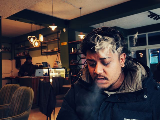 Young man exhaling vapor smoke indoors at a café, wearing a dark jacket, with warm pendant lights in the background.